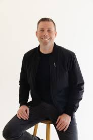 Ty Bennett, founder of Leadership Inc., sitting on a stool, smiling, wearing a black jacket and shirt, embodying the essence of a leadership expert and motivational speaker.
