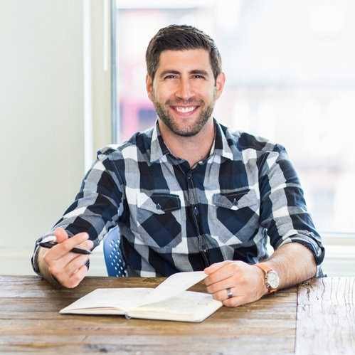 Keynote speaker and digital media strategist Jay Acunzo smiling at a desk with a notebook.